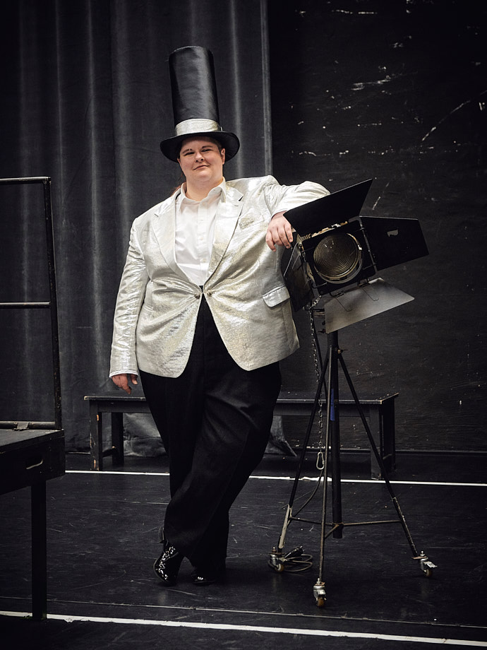 Die angehende Schauspielerin Nora Krohm stützt steht lässig auf den Bühnenscheinwerfer gestützt inmitten einer Probebühne. Sie trägt schwarze Schuhe und und eine schwarze Anzughose, eine weißes Oberhemd und ein silbrig glänzendes Jacket. Den Kopf ziert ein übermäßig hoher Zylinder.