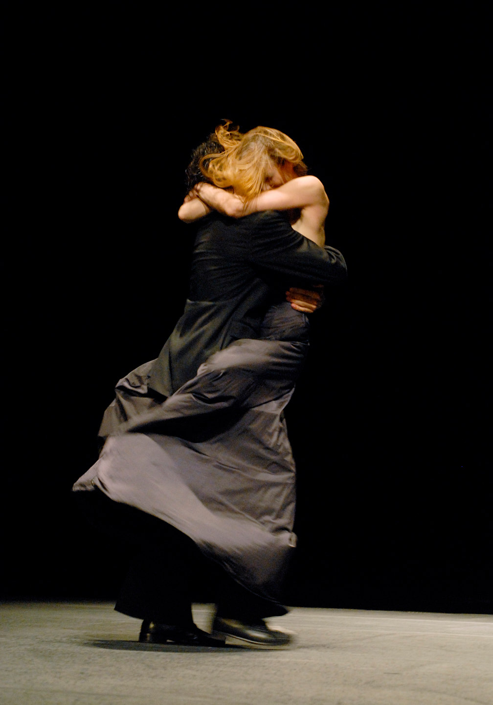 Szenenfoto aus einer Aufführung des Tanztheater Pina Bausch im Opernhaus Wuppertal. Ein tanzendes Paar, bei dem die Tänzerin die Beine um den Körper ihres Tanzpartners geschlungen hat, der über die Bühne schreitet.