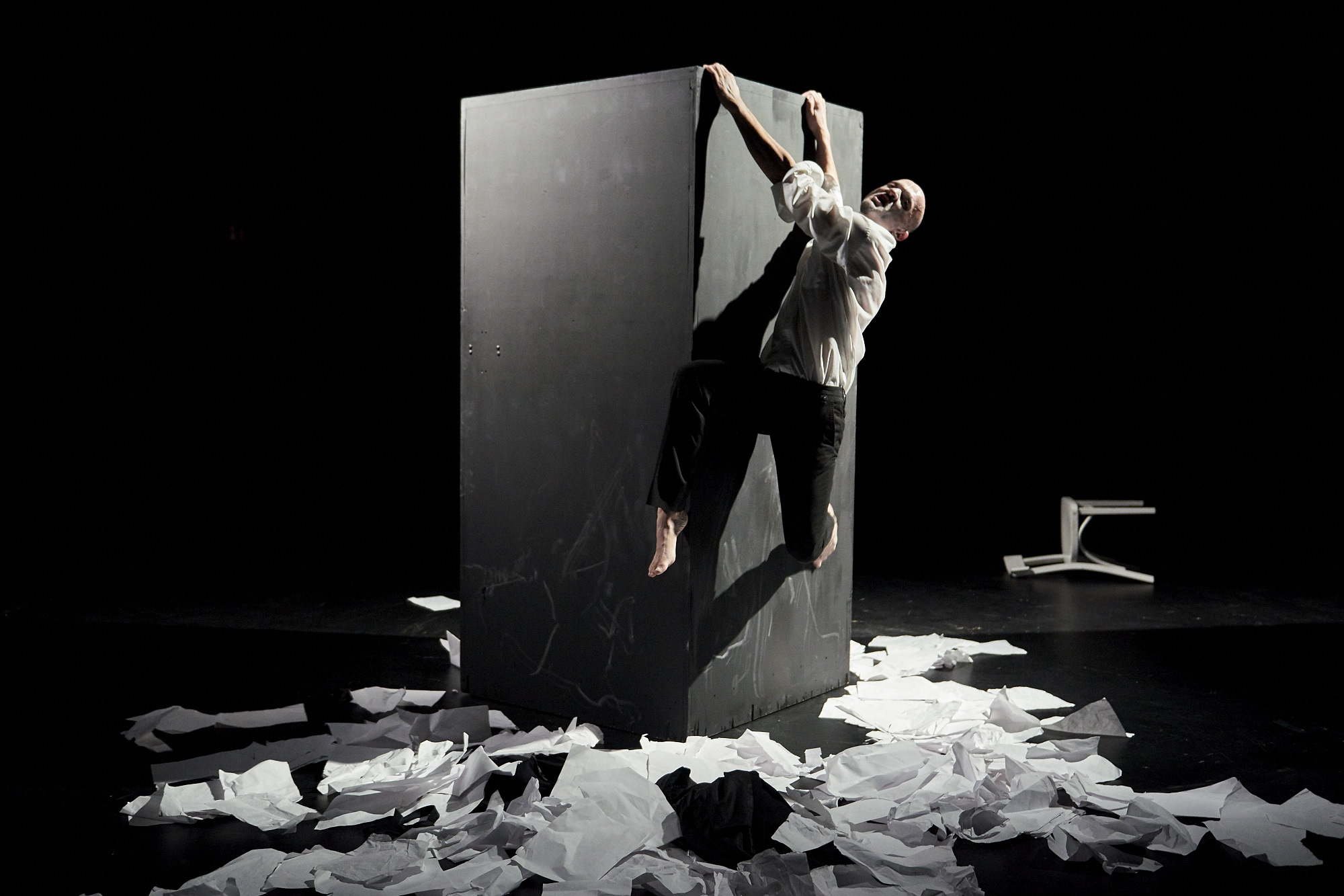 Theaterfotografie auf der sich ein Mann versuch nicht von einer Wand abzustürzen. Auf dem Boden ist ein Meer aus Papieren wild verteilt.